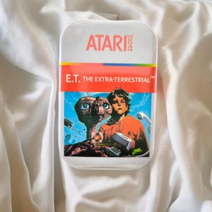 Könnte beinhalten: Eine rechteckige weiße Metalldose mit dem Atari 2600-Logo oben und dem Titel E.T. THE EXTRA-TERRESTRIAL in weißer Schrift auf rotem Grund. Die Mitte zeigt eine farbenfrohe Illustration der Figur E.T. und einer jungen Person.