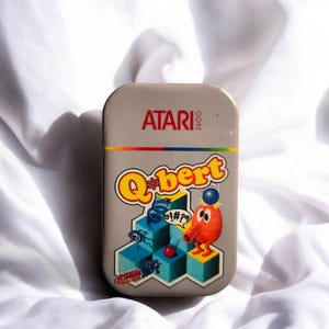 May include: A rectangular metal tin featuring the Atari 2600 logo at the top and the Q*bert video game title below. The graphic displays the orange Q*bert character on blue isometric cubes with a speech bubble containing symbols and small creatures.
