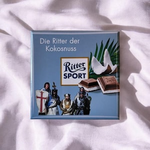 Könnte beinhalten: Eine quadratische Ritter Sport Schokoladenverpackung in Hellblau mit dem Text Die Ritter der Kokosnuss. Das Design zeigt eine Grafik von vier mittelalterlichen Rittern, zwei Schokoladenstücken mit Kokosfüllung und einem grünen Palmblatt.