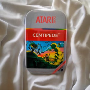Könnte beinhalten: Eine rechteckige silberne Metalldose mit dem Atari 2600 Logo oben. Ein rotes horizontales Band zeigt das Wort CENTIPEDE in weißer Schrift. Darunter befindet sich eine bunte Grafik mit einem gelb-grünen Tausendfüßler, einer Spinne und einer kleinen Figur.