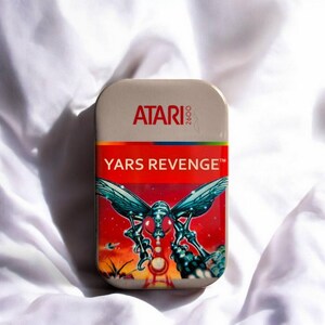 Könnte beinhalten: Eine rechteckige Metalldose mit dem roten Atari 2600-Logo oben und dem Schriftzug Yars Revenge in Weiß auf rotem Grund. Der untere Teil zeigt eine farbenfrohe Grafik eines insektenartigen Wesens vor einem roten Weltraumhintergrund.