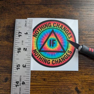 May include: A round sticker with the text "NOTHING CHANGES IF NOTHING CHANGES" in a rainbow tie-dye design. The sticker has a black triangle with the word "IF" inside. A ruler shows the sticker is approximately 3 inches in diameter.
