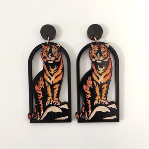 Könnte beinhalten: Ein Paar schwarze Holzohrringe mit einem Tigerdesign. Der Tiger ist orange und schwarz und sitzt auf einem Felsen. Die Ohrringe sind in Form eines Bogens.