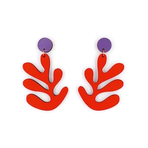 May include: A pair of red and purple earrings shaped like stylized leaves. The earrings are made of wood and have a modern, abstract design.
