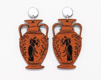 May include: A pair of orange and black ceramic vase earrings with a white background. The earrings are shaped like ancient Greek vases with a black silhouette of a woman on each vase.