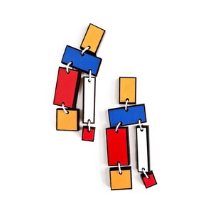 Pode incluir: Um par de brincos geométricos coloridos com quadrados e retângulos vermelhos, azuis, amarelos e brancos conectados por anéis prateados.