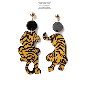 Pode incluir: Um par de brincos de madeira com um design de tigre. Os tigres são amarelos com listras pretas e estão sobre um fundo preto. Os brincos estão suspensos de ganchos dourados.