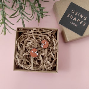 May include: A pair of fox-shaped wooden earrings in a brown cardboard box with brown paper shredding. The box has a black label that says "USING SHAPES ATHENS".