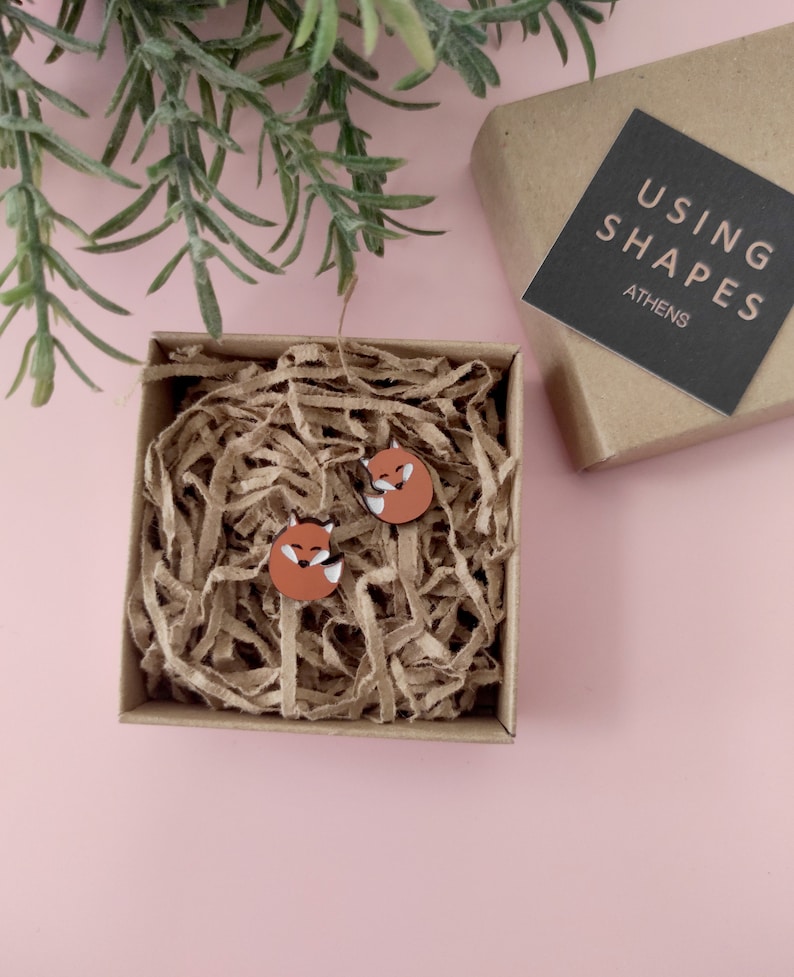 May include: A pair of fox-shaped wooden earrings in a brown cardboard box with brown paper shredding. The box has a black label that says "USING SHAPES ATHENS".