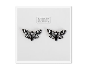 May include: A pair of black and white moth earrings with a skull design. The earrings are on a white background with the text "USINGSHAPES" in black.
