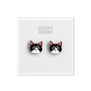 Op de afbeelding: Een paar zwarte en witte oorringen met een kattengezicht en roze oren. De oorringen zijn gemaakt door het bedrijf "Using Shapes".