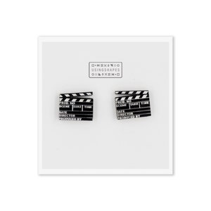 Op de afbeelding: Twee zwart-witte filmklapbord oorbellen. Elke oorbel heeft de tekst "PROD. NO", "SCENE", "TAKE", "TIME", "DATE", "DIRECTOR" en "PRODUCED BY". De oorbellen staan op een witte achtergrond met de tekst "USINGSHAPES".