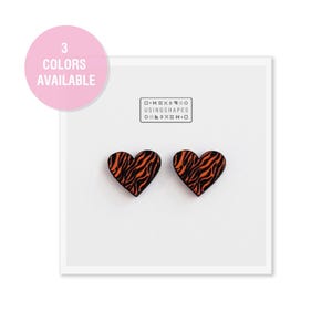 Pode incluir: Um par de brincos em forma de coração laranja e preto com um padrão de zebra. Os brincos são exibidos em um fundo branco com o texto "USING SHAPES" em preto.