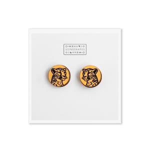 May include: A pair of yellow wooden earrings with a black cheetah print design. The earrings are round and have a smooth finish. The text "D.I.Y.W.O. USINGSHAPES O.I.D.I.M.O." is printed on a white card behind the earrings.