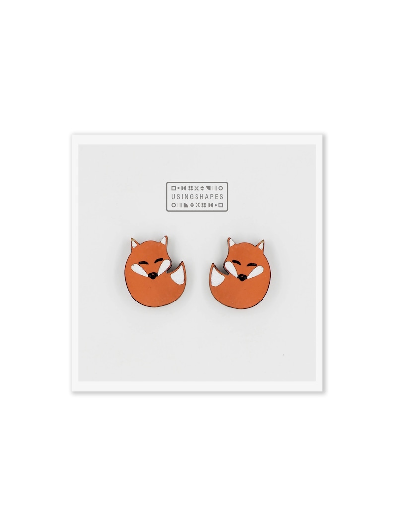 May include: A pair of orange fox-shaped earrings with white accents. The earrings are made of wood and have a simple, minimalist design. The text "USING SHAPES" is printed on a white background.