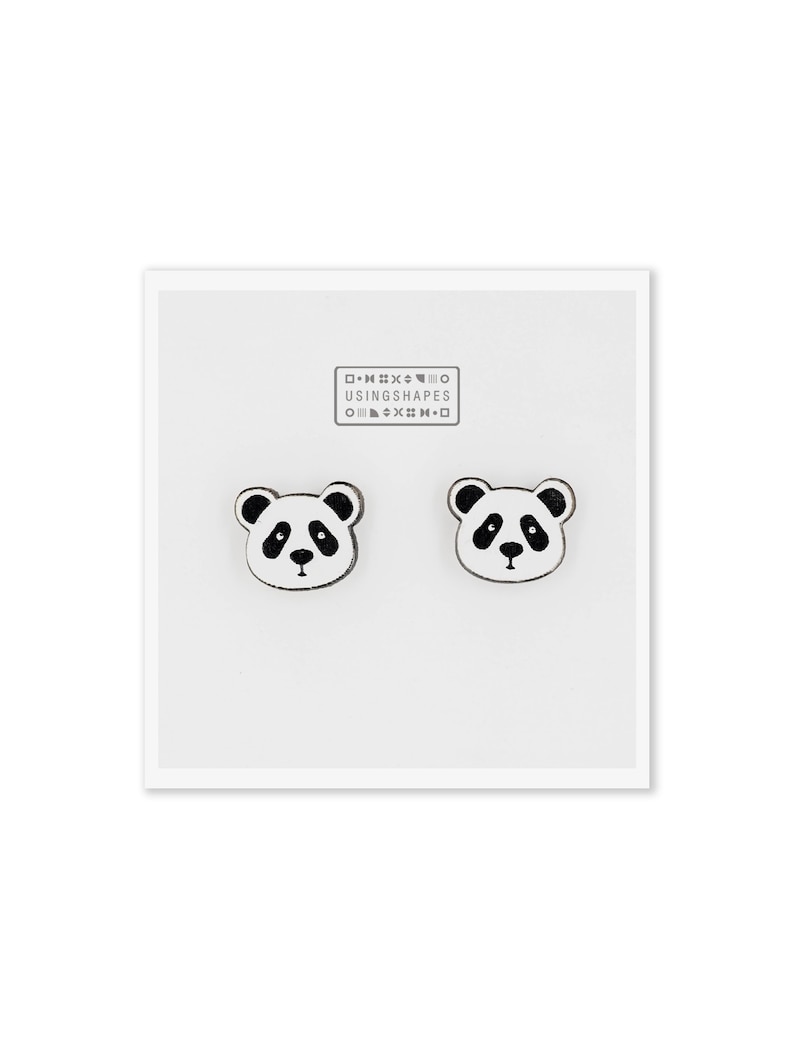 May include: A pair of white wooden panda bear earrings with black eyes, nose, and ears. The earrings are packaged on a white card with the text "USING SHAPES" printed in black.