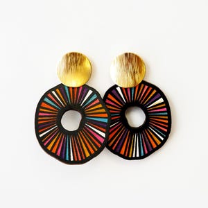 May include: A pair of colorful earrings with a black background and gold accents. The earrings are round and have a sunburst design with stripes of orange, pink, purple, blue, and white.