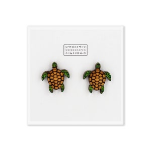 Könnte beinhalten: Ein Paar Holzohrringe in Schildkrötenform. Die Ohrringe sind braun mit grünen Akzenten und werden auf einer weißen Karte präsentiert. Die Karte hat ein Logo mit der Aufschrift "USINGSHAPES".