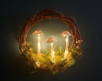 Può includere: Una ghirlanda fatta di ramoscelli con tre luci di fungo luminose. I funghi sono marroni e bianchi e sono circondati da muschio verde.