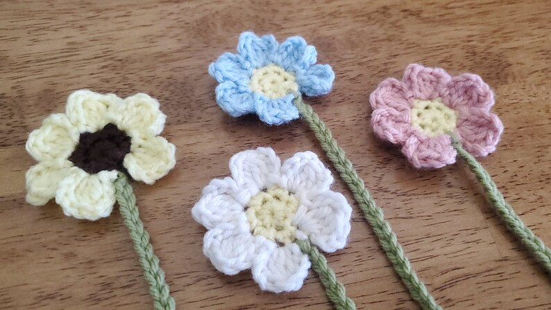 May include: Four crocheted flowers with green stems. The flowers are yellow, blue, white, and pink.