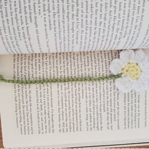 May include: A white crocheted daisy bookmark with a green stem and leaves. The bookmark is placed on an open book with text about the death of a character named Una.
