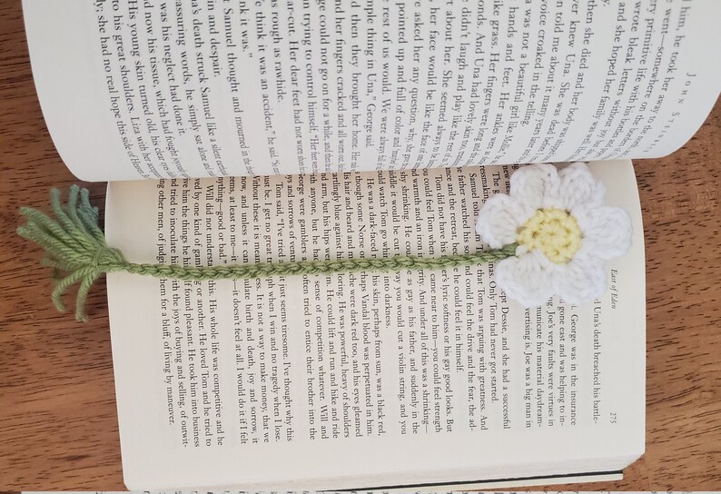 May include: A white crocheted daisy bookmark with a green stem and leaves. The bookmark is placed on an open book with text about the death of a character named Una.