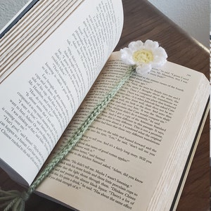 May include: A close-up of a book with a green crocheted bookmark. The bookmark features a white crocheted flower with a yellow centre. The book is open to a page with text about the story of Cain and Abel.