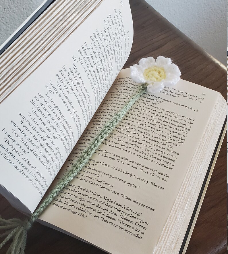 May include: A close-up of a book with a green crocheted bookmark. The bookmark features a white crocheted flower with a yellow centre. The book is open to a page with text about the story of Cain and Abel.