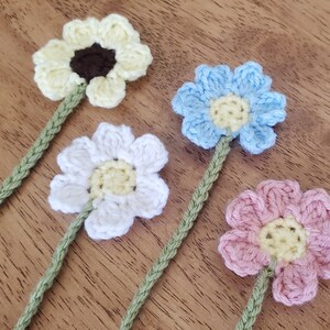 May include: Four crocheted flower stems made with green yarn. The flowers are yellow, white, light blue, and pink. Each flower has a yellow centre.