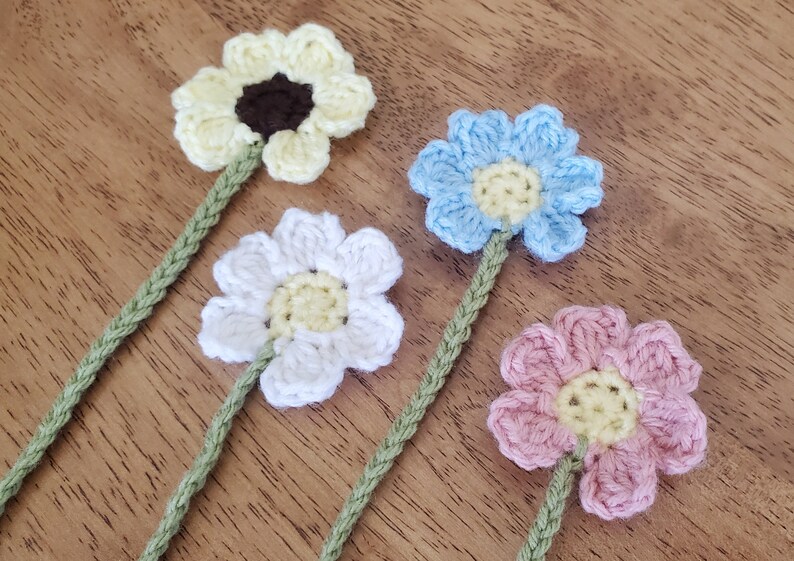 May include: Four crocheted flower stems made with green yarn. The flowers are yellow, white, light blue, and pink. Each flower has a yellow centre.