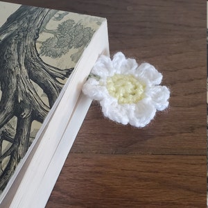 May include: A white crocheted daisy bookmark with a yellow centre. The bookmark is placed in the corner of a book.