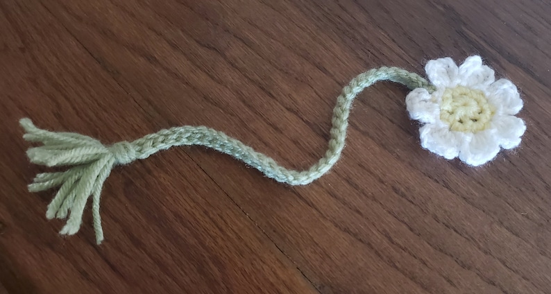 May include: A crocheted bookmark shaped like a daisy with a green stem and tassel. The flower is white with a yellow centre.