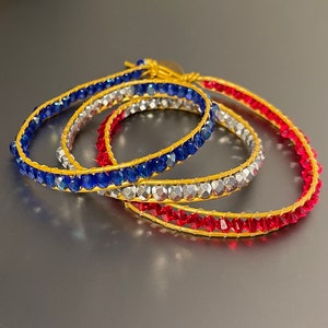 Puede incluir: Tres pulseras doradas con cuentas de cristal azul, plateado y rojo. Las pulseras están superpuestas.