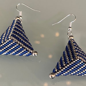 Puede incluir: Un par de pendientes hechos a mano con un diseño geométrico. Los pendientes son triangulares, con un patrón tejido de cuentas azules y plateadas. Tienen ganchos para las orejas plateados y pequeñas cuentas plateadas en las esquinas.