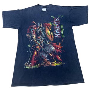 Puede incluir: Camiseta azul oscuro con un gráfico de un guerrero medieval sosteniendo un escudo y una espada. El guerrero está representado con una armadura roja, naranja y plateada. La palabra "Spawn" está impresa verticalmente en verde y blanco, con "Medieval" debajo.