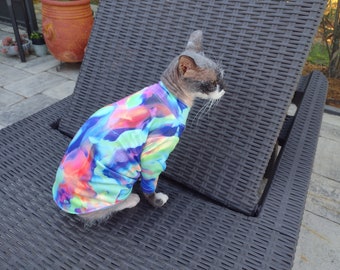 May include: A hairless cat wearing a colorful, abstract print shirt sits on a black wicker lounge chair.