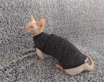 May include: A hairless cat wearing a black and grey sweater with a black trim. The cat is sitting on a white and grey fuzzy surface.