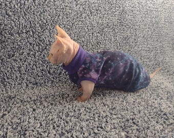May include: A Sphynx cat wearing a purple and blue galaxy-patterned sweater. The cat is hairless with pink skin and large ears. The sweater has a high collar and covers the cat's body. The cat is on a gray, textured surface.
