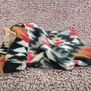 May include: A Sphynx cat peeks out from a colorful fleece blanket. The blanket features patterns in black, white, orange, and red. The cat's pink skin contrasts with the blanket's textures.