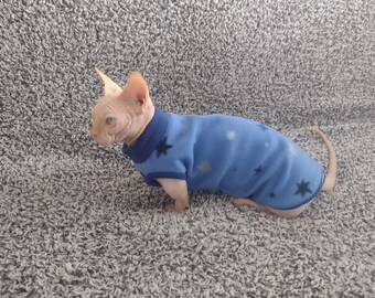 May include: A hairless cat wearing a blue fleece sweater with white stars. The sweater has a navy blue collar.