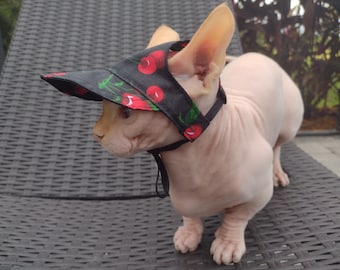May include: A hairless cat wearing a black cherry-patterned sun hat. The cat is sitting on a black wicker chair.