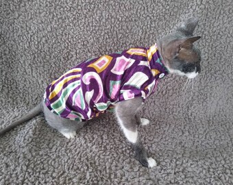 May include: A gray and white cat wearing a purple, pink, green, and gold patterned shirt. The shirt has a button closure at the neck.