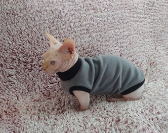 May include: A hairless cat wearing a gray and black sweater. The cat is sitting on a fluffy pink rug.