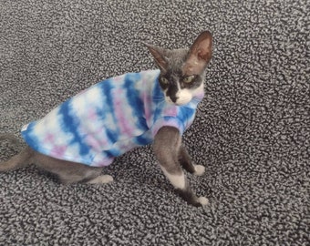 May include: A hairless cat wearing a blue, pink, and white tie-dye shirt. The cat is sitting on a gray carpet.