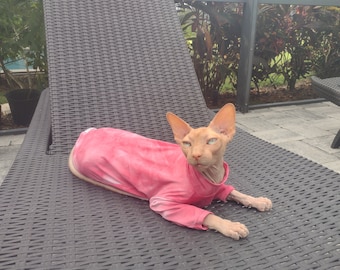 May include: A hairless cat wearing a pink tie-dye shirt is lying on a black wicker lounge chair.
