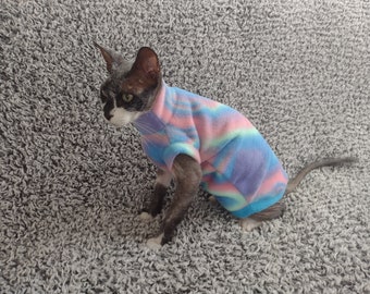 May include: A hairless cat wearing a colorful striped fleece sweater. The cat is sitting on a gray carpet.