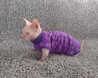 May include: A hairless cat wearing a purple sweater with white polka dots. The cat is sitting on a gray fuzzy surface.