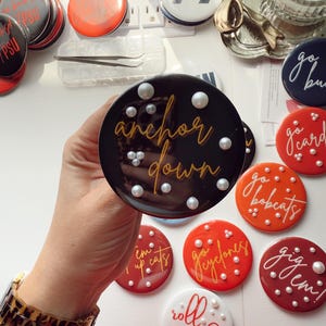May include: A collection of round, decorative buttons in various colors, including black, red, orange, and white. The buttons feature phrases like "anchor down," "go bulldogs," and "roll tide," along with pearl accents. A hand holds one button.