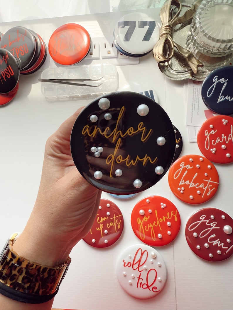 May include: A collection of round, decorative buttons in various colors, including black, red, orange, and white. The buttons feature phrases like "anchor down," "go bulldogs," and "roll tide," along with pearl accents. A hand holds one button.