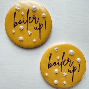 May include: Two round, mustard-yellow coasters with the phrase "boil er up!" in black script. Each coaster is adorned with faux pearls of varying sizes, adding a touch of elegance to the design. The coasters are placed on a white surface.